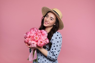 Photo of Beautiful young woman in straw hat with bouquet of peonies on pink background