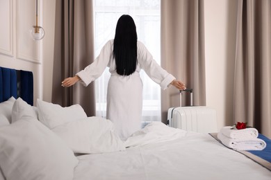 Young woman wearing bathrobe near window in hotel room, back view