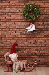 Christmas gnome on sleigh near brick wall with pair of ice skates and festive wreath indoors