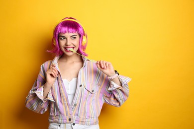 Fashionable young woman in colorful wig with headphones chewing bubblegum on yellow background, space for text