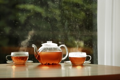 Teapot and cups of hot tea on wooden table against blurred background, space for text