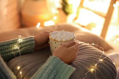 Woman holding cup of hot drink with marshmallows indoors, closeup. Magic Christmas atmosphere