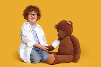 Photo of Little boy in medical uniform and toy bear with bandage on yellow background
