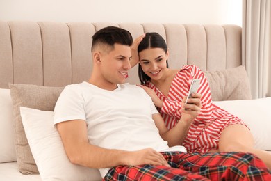 Photo of Happy couple in pyjamas using smartphone on bed at home