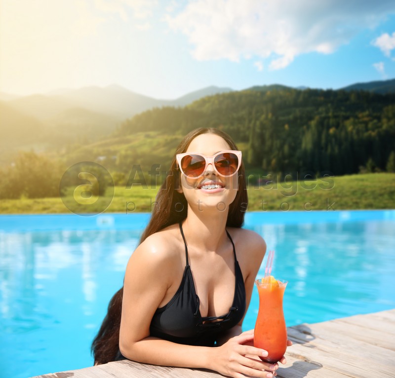 Beautiful young woman with cocktail in outdoor swimming pool at luxury resort and beautiful view of mountains on sunny day