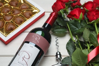 Photo of Bottle of red wine, beautiful roses and heart shaped chocolate candies on white wooden table, closeup