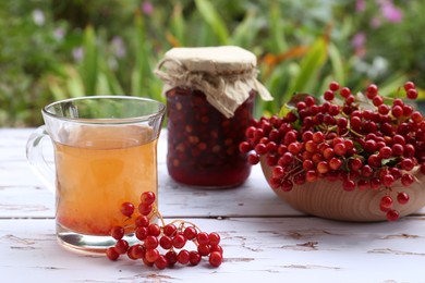 Photo of Cup of tea, jam and ripe viburnum berries on white wooden table outdoors