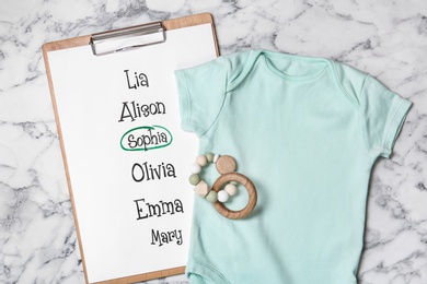 Bodysuit, list of baby names and toy on white marble background, flat lay
