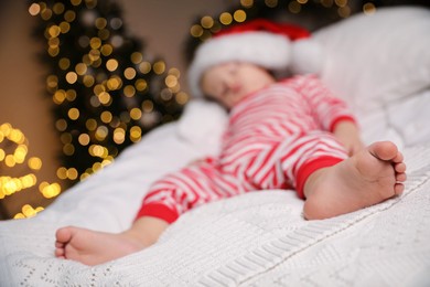 Photo of Baby in Christmas pajamas and Santa hat sleeping on bed indoors, focus on feet