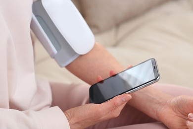 Woman checking blood pressure with modern monitor and smartphone indoors, closeup
