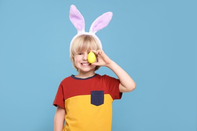 Photo of Happy boy in bunny ears headband holding painted Easter egg on turquoise background