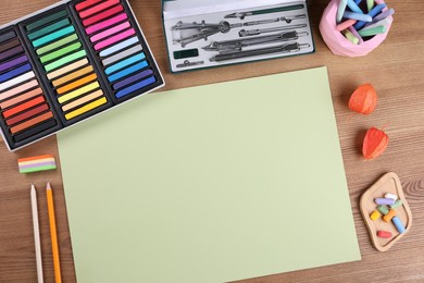 Blank sheet of paper, colorful chalk pastels and other drawing tools on wooden table, flat lay. Modern artist's workplace