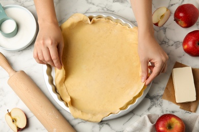 Woman putting dough for traditional English apple pie into baking dish at white marble table, top view