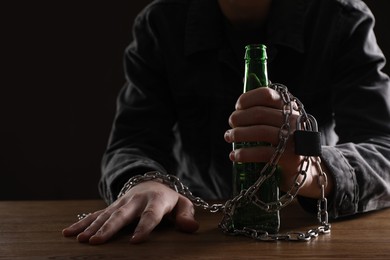 Alcohol addiction. Man chained with bottle of beer at wooden table, closeup