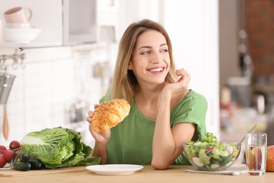 Photo of Woman eating croissant at table in kitchen. Diet failure