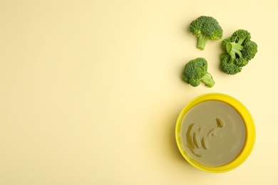 Baby food in bowl and fresh broccoli on yellow background, flat lay. Space for text