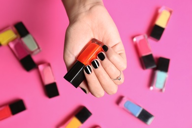 Woman with black manicure holding nail polish bottle on color background, top view