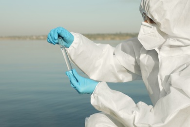 Scientist in chemical protective suit with test tube taking sample from river for analysis