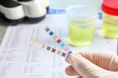 Doctor holding urine test strips near documents at table, closeup
