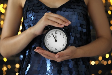 Woman holding alarm clock against blurred lights, closeup. New Year countdown