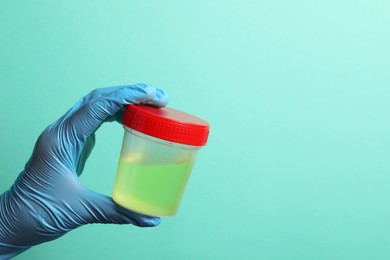 Doctor holding container with urine sample for analysis on turquoise background, closeup. Space for text