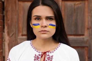 Photo of Young beautiful woman with drawings of Ukrainian flag on face near wooden door