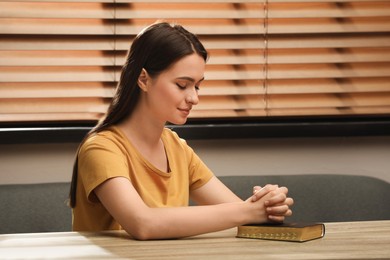 Religious young woman praying over Bible at wooden table indoors