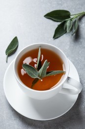 Cup of aromatic sage tea and fresh leaves on grey table