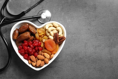 Heart shaped bowl with nuts and dried fruits near stethoscope on grey background, top view. Space for text
