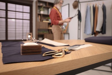 Photo of Professional tailor working in atelier, focus on table with scissors and pin cushion. Space for text