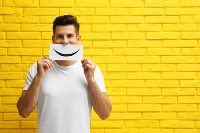 Man hiding emotions using card with drawn smile near yellow brick wall. Space for text