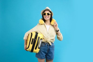 Happy female tourist with suitcase and travel pillow on light blue background
