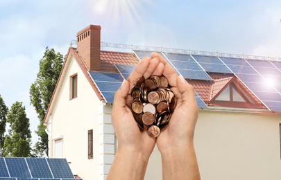 Woman holding coins against house with installed solar panels. Renewable energy and money saving