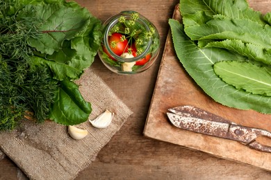 Fresh green herbs, jar with tomatoes, garlic and scissors on wooden table, flat lay