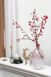 Hawthorn branches with red berries in vase, candles and deer figures on white table indoors