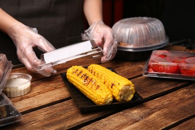 Waiter in gloves closing container with grilled corn cobs at wooden table, closeup. Food delivery service