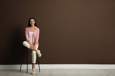 Beautiful young woman sitting on stool near brown wall. Space for text
