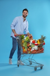 Young man with shopping cart full of groceries on light blue background