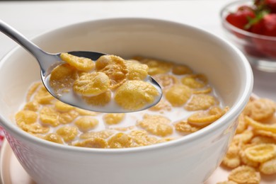 Spoon of delicious crispy corn flakes with milk above bowl on white table, closeup