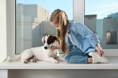 Cute little girl with her dog on window sill indoors. Childhood pet