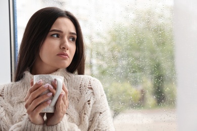 Sad beautiful woman with cup near window indoors on rainy day