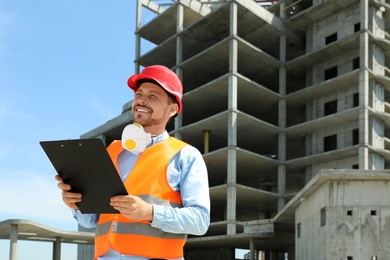 Professional engineer in safety equipment with clipboard at construction site. Space for text