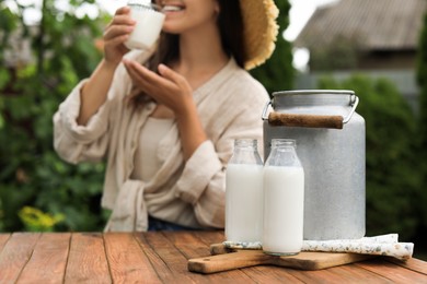 Photo of Smiling woman drinking milk at wooden table outdoors, focus on can and bottles