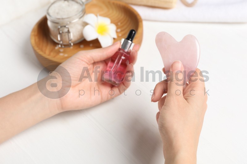 Photo of Woman holding gua sha tool and cosmetic at white wooden table, closeup