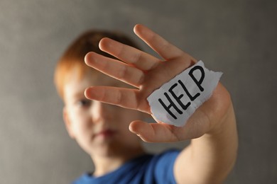 Photo of Little boy holding piece of paper with word Help against light grey background, focus on hand. Domestic violence concept