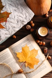 Photo of Book with autumn leaf as bookmark, acorns, scented candles and warm sweaters on wooden table, flat lay