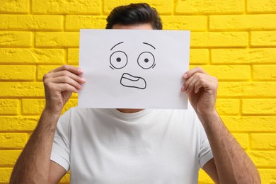 Man hiding emotions using card with drawn scared face near yellow brick wall
