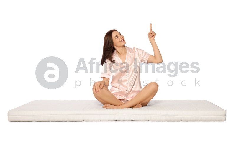 Young woman sitting on soft mattress and pointing upwards against white background