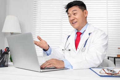 Happy doctor with laptop consulting patient at white desk in clinic. Online medicine