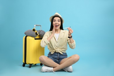 Happy female tourist with toy plane, suitcase and travel pillow on light blue background